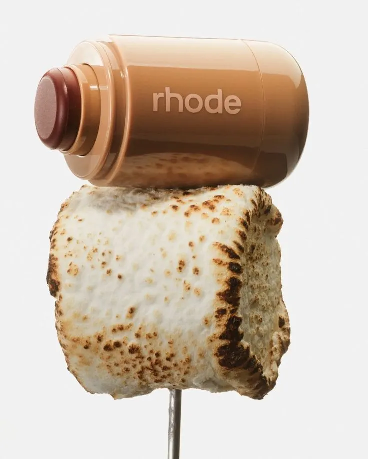 Produkt péče o pleť Rhode (balzám na rty, lip treatment) v hnědém obalu, ležící na opékaném marshmallow na špejli. Vizualizuje sladkou chuť a gurmánský zážitek v kosmetice.