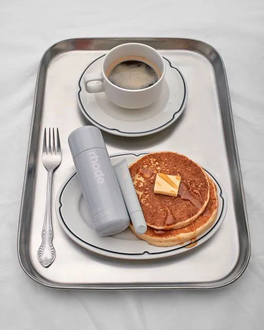 Ranní tác se snídaní: čerstvá káva, nadýchané palačinky s máslem a dva produkty Rhode (lahvička hydratačního krému a tuba balzámu na rty) na talíři. Evokuje pohodu, relaxaci a kvalitní ranní péči.